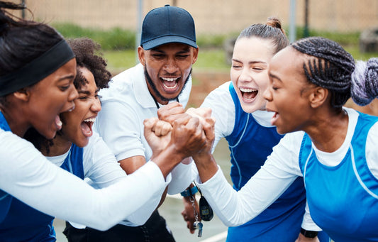A team of teen female athletes clasp hands with their male coach, cheering themselves on.